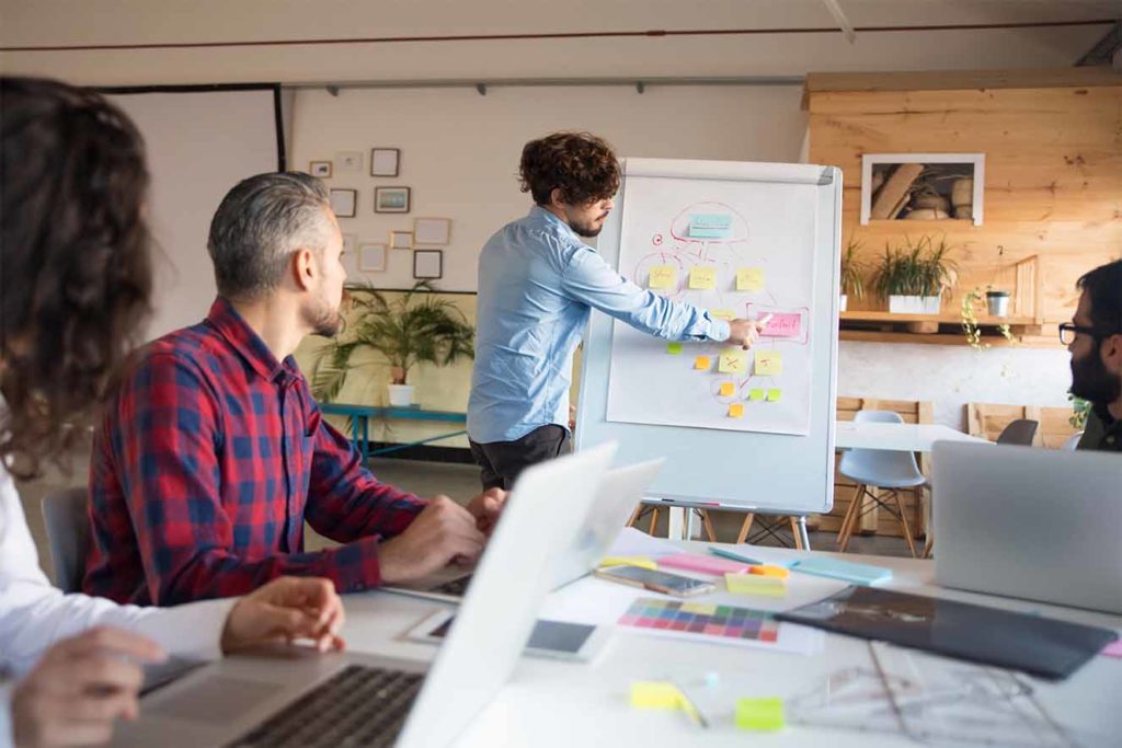 Creative group discussing ideas for project in boardroom, leader presenting chart on board. Business colleagues in casual working together in contemporary office space. Brainstorming concept