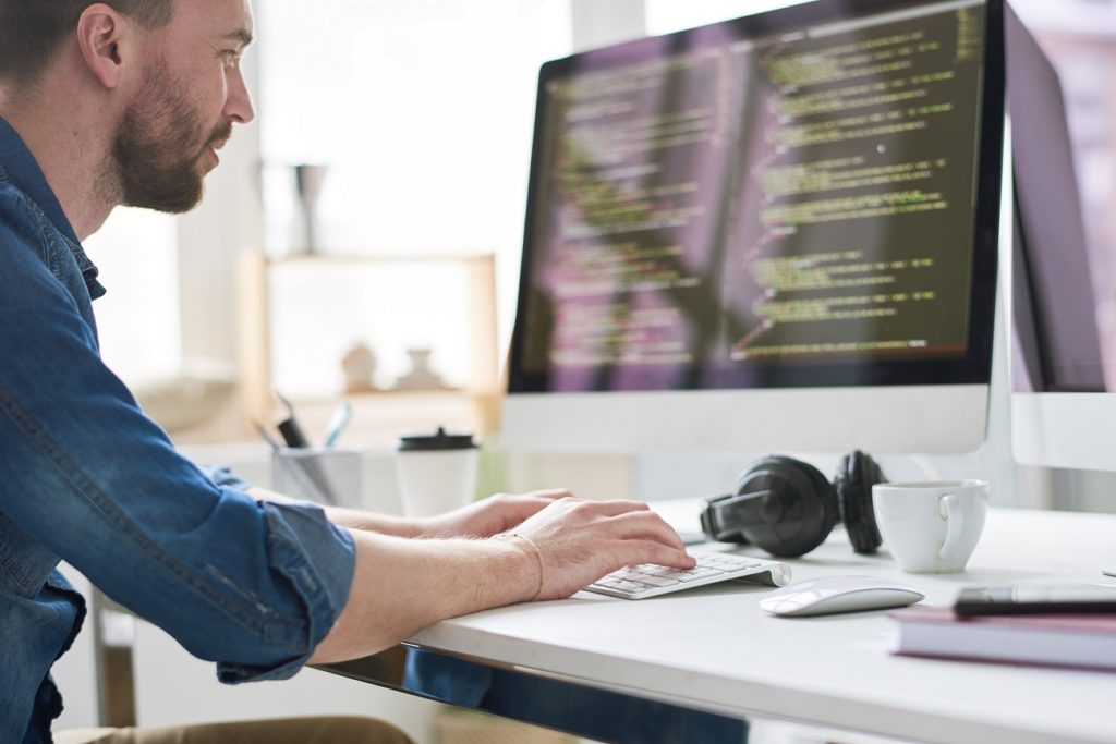 Serious pensive experienced male programmer typing code on computer while working on new software application in modern comfortable office