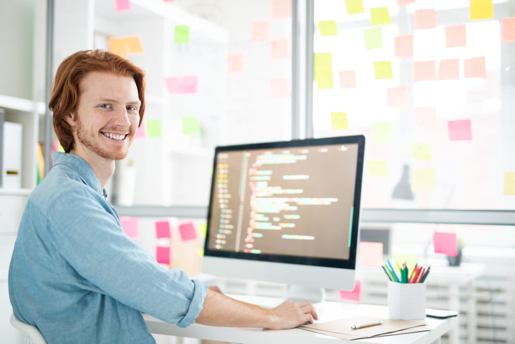 Young cheerful it-developer with toothy smile looking at you while sitting by workplace in front of computer screen