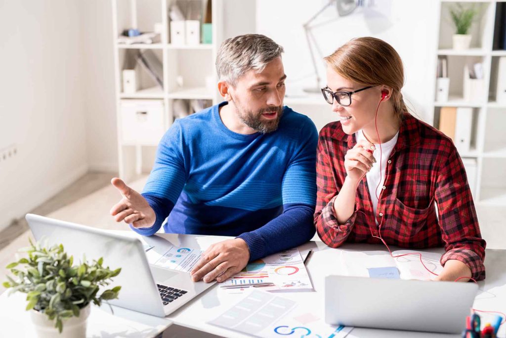 Cheerful optimistic young colleagues in casual clothing sitting at table and discussing marketing analysis in office: bearded man pointing at screen while explaining strategy to woman in earphones